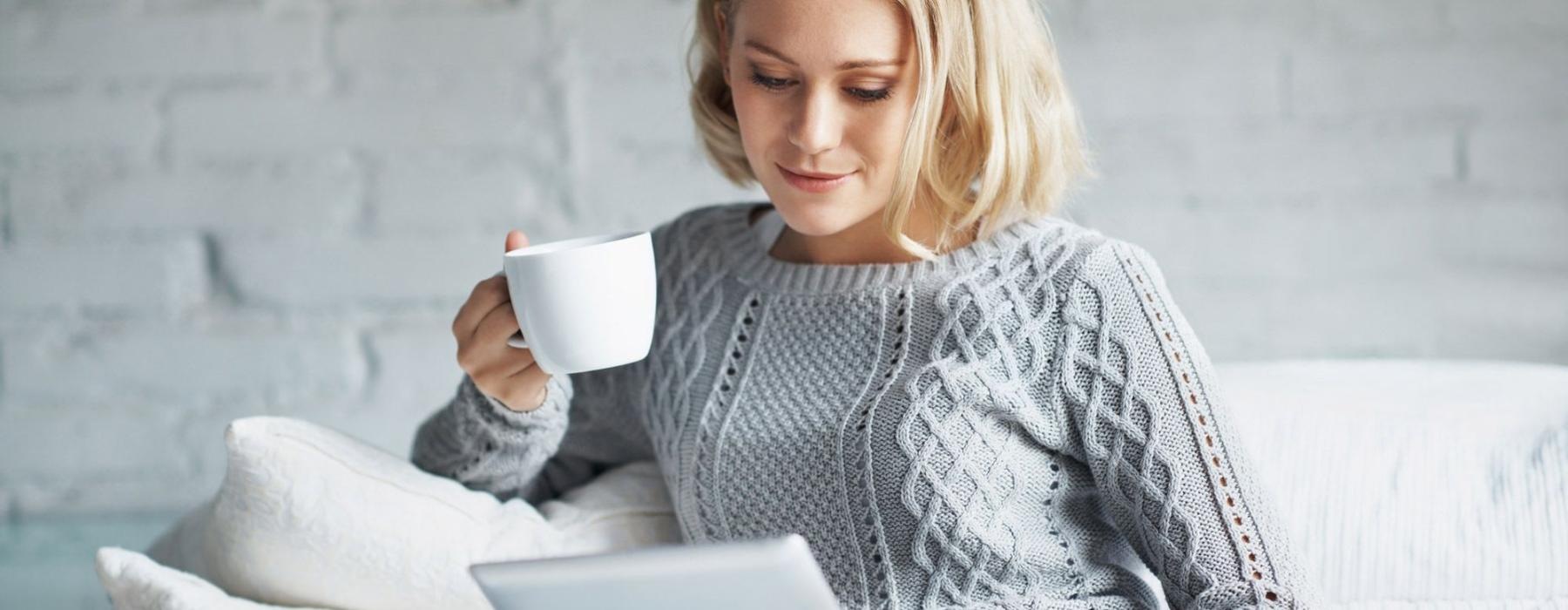 a woman sits on a couch with a cup of coffee and looks at her tablet